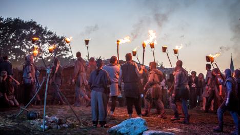 Mittelalterliche Menschen und Ritter mit Fackeln (von hinten) hinter abgebrannter Feuerstelle und zwei großen Steinen vor Holzhütten, im Hintergrund Baumkronen und Himmel bei Sonnenaufgang