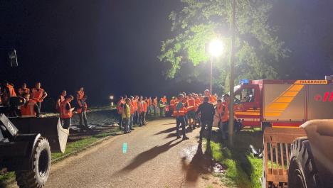 Personen mit orangen Warnwesten bei Nacht auf und neben Straße, rechts rote Feuerwehrfahrzeug, hinten Lichtstrahler vor Baum, links Ausschnitt von einem Bagger mit Schaufel