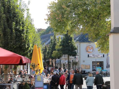 Brüggener Fußgängerzone mit Reihen von Bäumen und Gebäuden rechts und links, links offene rote und geschlossene gelbe Sonnenschirme, darunter teilweise besetzte Tische und Stühle, entlang der Straße verschiedene Menschen 