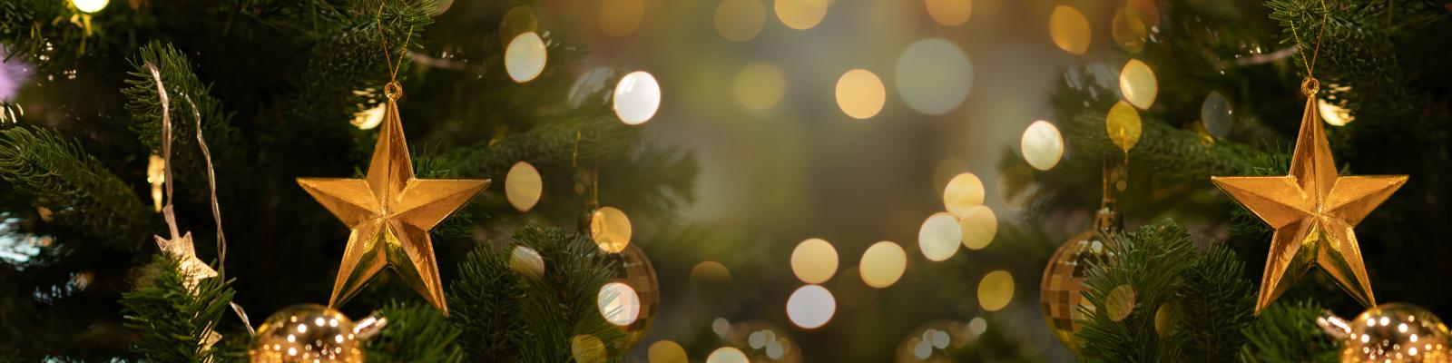 Nahaufnahme von einem grünen Tannenbaum geschmückt mit goldenen Sternen, goldenen Kugeln und Lichterketten, die sternförmige oder runde Leuchten haben, im Hintergrund mittig verschwommene Lichter