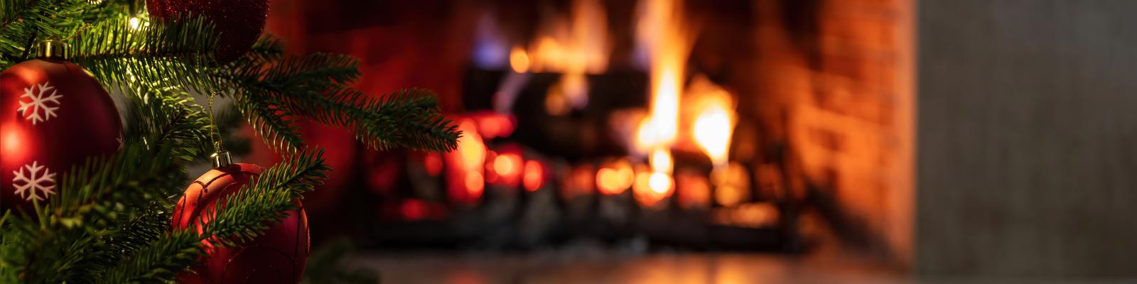 Links kleiner Ausschnitt von grünem Tannenbaum geschmückt mit roten und goldenen Kugeln, teils mit Muster oder Glitzer, im Vordergrund unten leerer Holztisch, im Hintergrund verschwommen Blick auf brennende Feuerstelle 
