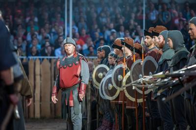Ritter mit rot-grauer Kleidung links von einer Reihe von weiteren Ritten mit Speeren und Schildern vor Zuschauern auf einer Tribüne verkleidet mit Holzlatten