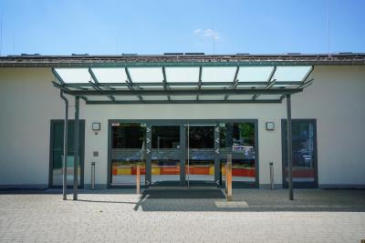 Überdachte Eingangstür aus Glas zur Burggemeindehalle Brüggen, links und rechts zwei weitere Eingangstüren, weiße Fassade, Spiegelbild von bunten Sitzgelegenheiten in Scheiben, vor der Eingangstür zwei silberne Pfeiler und hellgrau gepflasterter Fläche, über dem Dach blauer Himmel