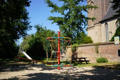 Buntes Klettergerüst, dahinter Wippe und Metallrutsche in Sandgrube, rechts daneben hölzerne Sitzbank mit Tisch auf grünem Rasenstück, rechts davon steinige Kirchenmauer und Borner Kirche, vor Kirchenmauer Spielplatzschild an Metallstange, Im Hintergrund dichter Baumbewuchs 