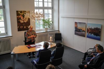 Trauung im Trauzimmer des Rathauses: Stehende Frau hinter hölzernen Tisch mit silbernen Beinen, auf Tisch Kerze und Papiere in roter Mappe, vor Tisch zwei sitzende Personen auf Stühlen, im Hintergrund Fenster mit Fensterbank und Heizkörpern darunter, Bild an weißer Wand dazwischen, rechts an weißer Wand zwei Bilder, unten und rechts Ausschnitte von sitzenden Personen auf Stühlen