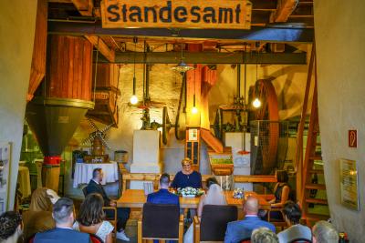 Trauung im Standesamt Brachter Mühle: sitzendes Brautpaar am hölzernen, dekorierten Tisch, links und rechts vom Tisch je eine sitzende Person, hinterm Tisch sitzende Person, oben hözernes Schild mit Aufschrift "Standesamt", im Hintergrund Innenausstattung und hölzerne Teile der Mühle, unten im Bild sitzende Menschen von hinten