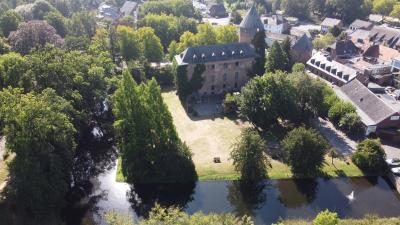 Panoramabild von Burg Brüggen mit teils trockener Burgwiese, Burgweiher und dichtem, teils vereinzelten Baumbewuchs, rechte obere Ecke mehrere Gebäude