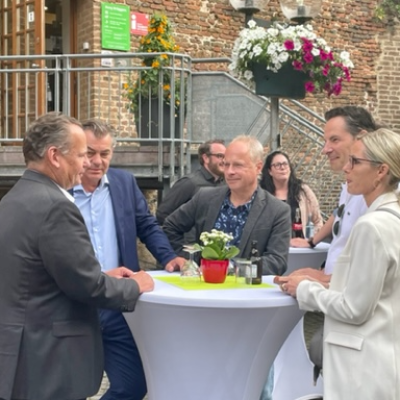 Menschen in förmlicher Kleidung stehend an dekorierten Stehtischen mit weißen Hussen, grünem Tischset mit Blume in rotem Topf und Getränken vor einer Laterne mit Blumenampel und Metalltreppe zur Eingangstür der Touristinformation in der Burg