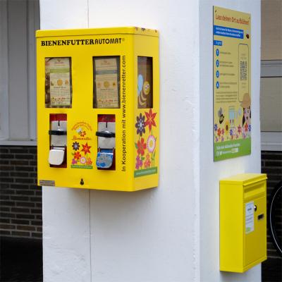 Gelber Automat mit bunten Blumen, Münzfach, Bienen und Informationstext an weißer Säule vor Gebäude, gelber Briefkasten mit Informationen und Plakat darüber an rechten Seite der Säule