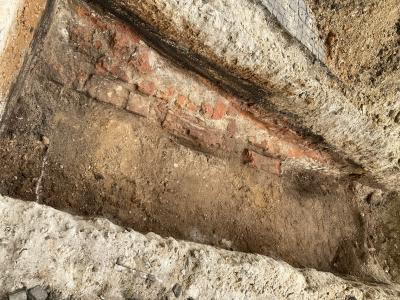 Rechteckige Ausgrabung mit Erde und Stück Mauer aus roten Klinkersteinen im Loch, rechts und links erste Erdschicht unter der Pflasterschicht, links Ausschnitt vom Pflaster und Hügel mit Erdböden aus dem Loch