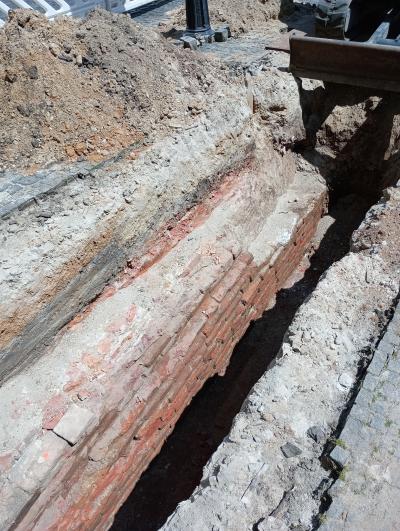 Rechteckige Ausgrabung angestrahlt von Sonne mit Erde und links Stück Mauer aus roten Klinkersteinen im Loch, rechts und links erste Erdschicht unter der Pflasterschicht, rechts und links daneben Teil vom Pflaster, links Hügel mit Erdböden aus dem Loch, im Hintergrund Ausschnitt weißer Absperrung, unterer Teil vom Laternenpfahl und Ausrüstung für Grabung, seitliche Ansicht