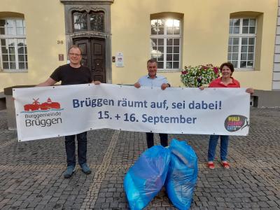 Drei Personen halten weißes Banner auf gepflasterten Boden vor gelben Gebäude mit drei Fenstern, und brauner Eingangstür: links Logo der Burggemeinde Brüggen, mittig blauer Schriftzug „Brüggen räumt auf, sei dabei!, 15. + 16. September“, unten rechts Logo World Cleanup Day, Mann links mit Brille, schwarzem T-Shirt, dunkel blauer Jeans und blauen Schuhen, Mann in der Mitte mit hellblauem Hemd, davor zwei befüllte, blaue Müllsäcke, Frau rechts mit roter Bluse, blauer Jeans und roten Sandaletten