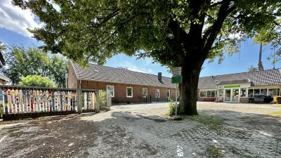 Große gepflasterte Fläche mit Gras dazwischen, mittig großer Baum mit rindigen Baumstamm und Baumkrone mit grünen Blätter, vor Baum weißes und grünes Schilde an Metallstange, links Metallzaun mit zwei Toren versehen mit bunt bemalten Latten, im Hintergrund L-förmiges Gebäude mit weißen Fensterahmen, rechts Eingangstür, datüber grünes Schild mit gelber Schrift "Kindergarten Lüttelbracht", rechts vor Gebäude Mülltonnen, links vom Gebäude grüne Bäume, hellblauer, leicht bewölkter Himmel