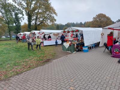 im Mittelpunkt 6 Stände mit weißen Abdeckungen und unterschiedlichen Verkaufsartikeln auf Weihnachtsmarkt am Weißen Stein, zwischen den Ständen einzelne Personen, vorne grau gepflastert, links davon grüne Wiese, im Hintergrund dichter Baumbewuchs, grauer Himmel 