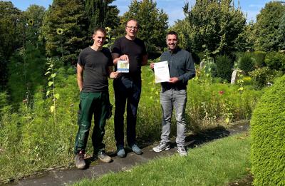Drei Männer stehend in Reihe auf gepflasterten Gehwegsplatten vor dichtem, grünem Wiesen-, Strauch- und Baumbewuchs, unten rechts grünes Stück Rasen und Ausschnitt von einem Busch, Mann links mit schwarzem T-Shirt, grüner Arbeitshose und schwarzen Arbeitsschuhen und Mann in der Mitte mit Brille, schwarzem T-Shirt, blauer Jeans und blau-grauen Schuhen halten zusammen Auszeichnung in Händen, Mann rechts mit dunkelgrauem Hemd, hellgrauer Hose und Schuhen hält Urkunde in den Händen