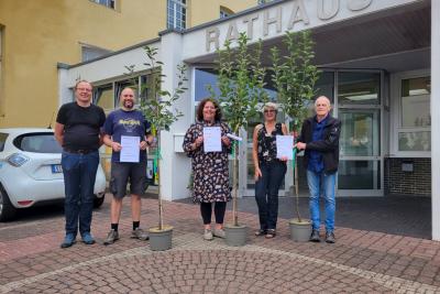 5 Personen auf rot-grau gepflasterten Platz vor Eingang des gelb-weißen Rathauses, von links nach rechts: Mann mit Brille, schwarzem T-Shirt, Jeans, blau-grauen Schuhen, Mann mit Glatze blauem T-Shirt, kurzer Hose, grauen Schuhen, Frau mit braunen Locken, Blumenkleid, schwarzer Leggins, grauen Schuhen, Frau mit kurzen Haaren, Brille, Bluse, Jeans, Sandalen, Mann mit schwarzer Lederjacke, blauem Hemd, Jeans, grauen Schuhen, drei Personen mit Urkunden in Händen, zwischen Personen kleine Bäume in grauen Töpfen