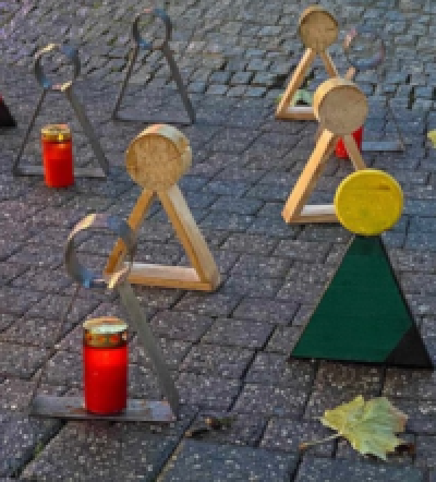 Selbstgemachte Figuren aus Holz oder Metall, teils bunt bemalt, Form unten bestehend aus Dreieckprisma, teils gefüllt, sonst nur Rahmen und oben auf Spitze kreisförmiges Prisma, teils gefühlt, sonst nur Rahmen, bei einzelnen Figuren Kerzen im roten Glas mit goldenen Deckel, Figuren stehen auf gepflasterten Boden, vorne rechts grünes Blatt
