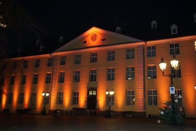 Großes Gebäude mit vielen Fenster wird mit orangen Lichtstrahlern beleuchtet, mittig unten Eingangstür mit Treppenaufgang und oben Wappen von Brüggen, vor Gebäude gepflasterter Platz mit drei leuchtenden Laternen mit je drei Leuchteinheiten, an Laternen Tannenbäume befestigt, Aufnahme bei Nacht