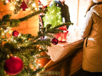 Im Vordergrund links Ausschnitt von Tannenbaum geschmückt mit Lichterkette, roten Weihnachtskugeln und Tannenbäumen, im Hintergrund Ausschnitt von Weihnachtsmarkthütte aus Holz mit Theke, Theke mit Tischdecke, Weihnachtsdeko, in und vor der Hütte je eine Person mit Winterjacke, Person vor Hütte mit beigen Wintermantel und Mütze