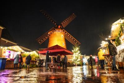 Bildaufnahme vom Boden von angestrahlter Brachter Mühle mit vier Flügeln, im Vordergrund nasser Platz vor Brachter Mühle mit einzelnen Menschen in dicker Winterkleidung, links und rechts beleuchtete Weihnachtsmarkthütten aus Holz, mittig roter Schrim, rechts davon grüner Tannenbaum mit Lichterkette, dunkler Himmel bei Nacht