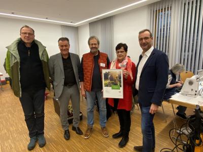5 Personen im Halbkreis, Holzboden, weiße Wände & Decke, Fenster bedeckt von Rollos, von links nach rechts: Mann mit Brille, grüne Jacke, Pullover, Jeans, blau-grauen Schuhen, Mann mit grauen Anzug, schwarzen Hemd & Schuhe, Mann mit Brille, oranger Weste, Pullover, Jeans, braune Schuhe, Frau mit Brille, rote Strickjacke, Schal, schwarze Hose & Schuhe hält Bilderrahmen mit Urkunde, Mann mit Brille, blauem Sacko, weißem Hemd, Jeans, im Hintergrund Tische & Stühle, wo Menschen Sachen mit Werkzeugen reparieren