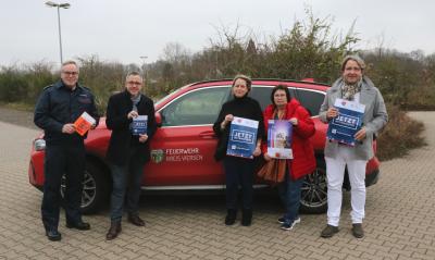 5 Personen halten Plakate vor rotem Auto mit Wappen Kreis Viersen & weißer Schirft "Feuerwehr Kreis Viersen", von links nach rechts: Mann mit Brille, dunkler Feuerwehr-Uniform, Mann mit Brille, grauem Schal, schwarzen Mantel, dunkle Hose & Schuhen, Frau mit schwarzem Oberteil & Schuhe, Jeans, Frau mit Brille, rote Jacke, Schal, Jeans, Turnschuhen, Mann mit grauem Mantel, Schal, weiße Hose, graue Schuhen, gepflasterter Boden, im Hintergrund kahler Strauch- & Baumbewuchs, Straßenlaterne, grau bewölkter Himmel