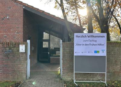 Rechts Weißes Schild an rollbarer Pinnwand auf Wiese mit Blättern vor Mauer mit roten Backsteinen, auf Plakat in schwarzer Schrift „Herzlich Willkommen zum Fachtag „Väter in den Frühen Hilfen““, darunter Logos der Beteiligten u.a. Kreis Viersen, links auch Mauer und Wiese mit Blättern, dazwischen gepflasterter Weg zum überdachten Eingang eines Gebäudes mit roten Backsteinen, rechts hinter Mauer Bäume mit einzelnen Blätter, Im Hintergrund Sonne