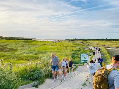 Panoramabild über einen Weg zum Strand und Meer: im Vordergrund unten gehen Menschen in Sommerkleidung Steintreppen links hoch und rechts runter, mittig flacher Aufstieg für Rollstuhlfahrende, unten an der Treppe steht Schild und verläuft Weg aus Holzbohlen, der zum Strand führt, Menschen auf dem Weg, links und rechts vom Weg grüne Dünenlandschaft mit Gras, im Hintergrund Sandstrand und Meer mit Personen, Himmel blau mit weißen Wolken