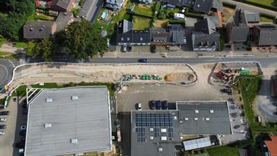 Luftaufnahme von oben (Vogelperspektive): unten zwei Gebäude mit Sicht auf Dächer, unten links und vor rechten Gebäude Parkplätze mit Autos, rechts unten grüner Pflanzenbewuchs, mittig zwei Baustellenbereiche mit Geräten, Boden bestehend aus Sand und Erde, eingerahmt durch Schrankenzaun, oben Wohngebiet mit verschiedenen Häusern, dazwischen Grünflächen und grüner Baumbewuchs