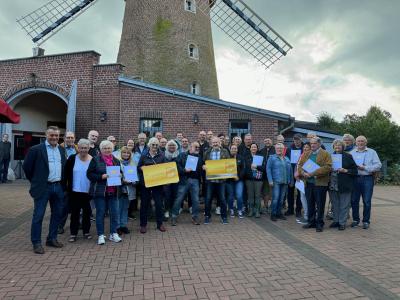 Stehende Gruppe von unterschiedlich alten und großen Menschen in bunter Kleidung mit Urkunden in den Händen auf gepflasterten Platz, 2 Personen halten je eine goldene Ehrenamtskarte vor Körper, im Hintergrund Gebäude der Brachter Mühle aus rotem Klinker, rechts mit schrägen Dach, links mit offener großer Tür, dahinter Mühlenturm bis zum dritten Fenster sichtbar und unterer Teil von 2 Mühlenflügeln, hinten rechts dichter grüner Strauchbewuchs, grau bewölkter Himmel