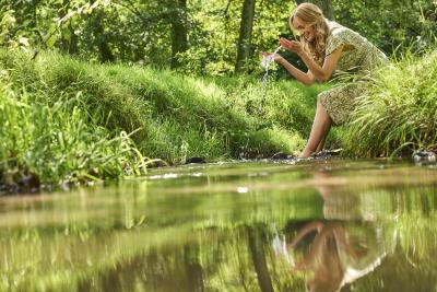 Im Vordergrund Nahaufnahme von Fluss oder Bach, links und rechts Ufer mit dichtem grünen Grasbewuchs, rechts sitzt Frau am Ufer mit gemusterten Kleid, blonden Haaren und Füßen im Fluss, sie lässt Wasser aus Händen fallen, die Hände hält sie vor sich auseinander, im Hintergrund dichter grüner Strauch- und Baumbewuchs, Frau, Ufer und Bäume spiegeln sich im Wasser