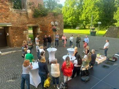 Aufnahme von schräg oben: Gruppe von bunt gekleideten Menschen verschiedenen Alters stehen auf grau gepflasterten Platz mit Stehtischen, die weiße Hussen und Deko haben, rechts unten Ausschnitt einer Bühne und kleines Buffet, auf Platz zwei Laternen, im Hintergrund Teile der Burgmauer, teils grün bewachsen, links Holztür, hinter Burgmauer grüne Wiese und dichter grüner Baumbewuchs