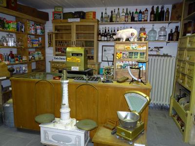 Tante Emma Laden: mittig Holztresen mit grüner altmodischen Kasse und rechts Gewürzregal mit Gewürzen, darauf weißen Sparschwein, vor Tresen zwei Waagen, links und rechts Holzregale gefüllt mit diversen Essen und Trinken, hinter Tresen links Anrichte aus Holz mit Glastüren oben, nur teils befüllt mit Flaschen, rechts hinterm Tresen zwei Holzbretter als Hängeregale mit verschiedenen Flaschen drauf, darunter weißer Heizkörper
