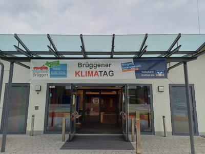 Eingang Burggemeindehalle Brüggen: Eingangstür bestehend aus zwei Flügeltüren aus Glas stehen offen, Blick in die Halle auf geschlossene Glastür, davor Teppichleiste und silberne Standsäulen, links und rechts von der Tür Lampen und zwei bodentiefe Fenster mit Milchglas, darüber Vordach aus Glas und Metallgerüst, daran hängt Banner zum Klimatag, links auf Banner Logo Klimatag, mittig Schriftzug Klimatag und rechts Volksbank Logo, oben bewölkter Himmel