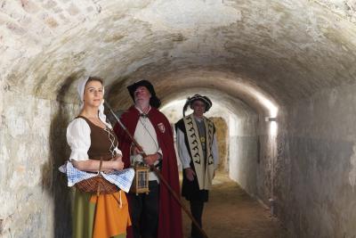 3 Personen in mittelalterlicher Kleidung stehen in Tunnel, helle, alte Wände, brauner, erdiger Boden, Personen von links nach rechts: Frau mit weißer Haube, braunen Oberteil mit weißen Ärmeln, grüner Rock, orange Schürze, brauner, geflochtener Holzkorb über rechten Arm mit blau-weiß karierten Handtuch, Mann mit schwarzen Hut, weißen Bart & Hemd, roten Mantel, in Händen Speer & Laterne, Mann mit dunklem Hut & Feder, weißem Hemd, langer dunkler Stoffweste, heller Schal über Schultern mit dunklem Muster