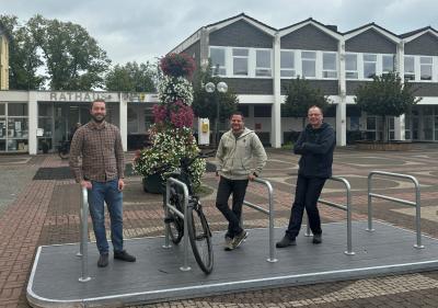Fahrradflunder: Plattform mit 5 aufgebauten Anlehnbügeln, schwarzes Fahrrad lehnt an zweiten Anlehnbügel von links, 3 Männer stehen an Anlehnbügeln (von links nach rechts): Mann mit Bart, karriertem Hemd, Jeans & schwarzen Schuhen, Mann mit heller Jacke, schwarzer Hose & grünen Schuhen, Mann mit Brille, dunkelblauer Jacke, schwarzer Hose & Schuhe, Flunder steht auf gepflasterten Platz, dahinter großer Blumenkübel & drei Bäume, im Hintergrund weiß, graues Gebäude, Baumkronen & grau bewölkter Himmel