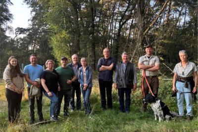 10 Menschen stehen auf grüner Rasenfläche in Reihe nebeneinander für Gruppenbild, rechts sitzt schwarz-weißer Hund vor Menschen, im Hintergrund dichter grüner und holziger Strauch- und Baumbewuchs bei Sonnenschein