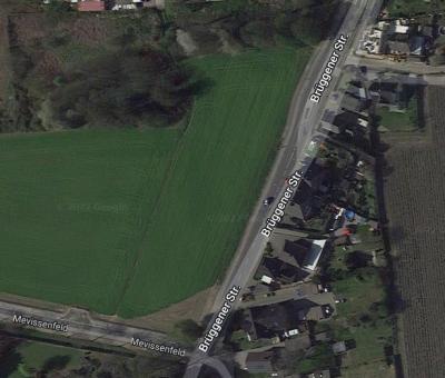 Luftaufnahme von grünen Feld, rechts und unten vom Feld Straßen, Straßen grau mit weißer Beschriftung der Straßennahmen, oben links dichter grüner Baumbewuchs, rechts und unten Gebäude von oben, Grünflächen und brauner Acker 