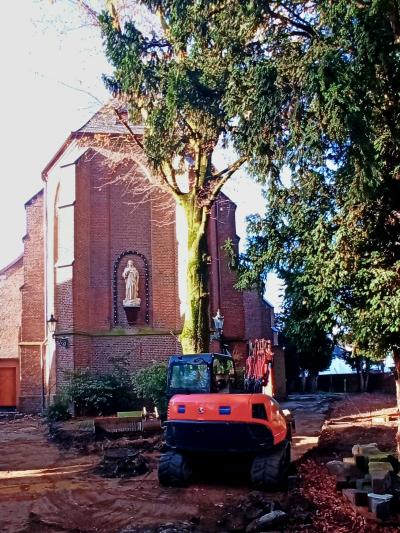 Kleiner schwarz-roter Bagger steht auf brauner Erdfläche, rechts liegen einzelne Steine, hinter Bagger Baum mit grünen Blättern und Sträucher vor Gebäude mit rotem Klinker, an Gebäudewand helle Kirchenstatue, Gebäude ist Kirche in Bracht, rechts und im Hintergrund dichter grüner Baumbewuchs, heller Himmel