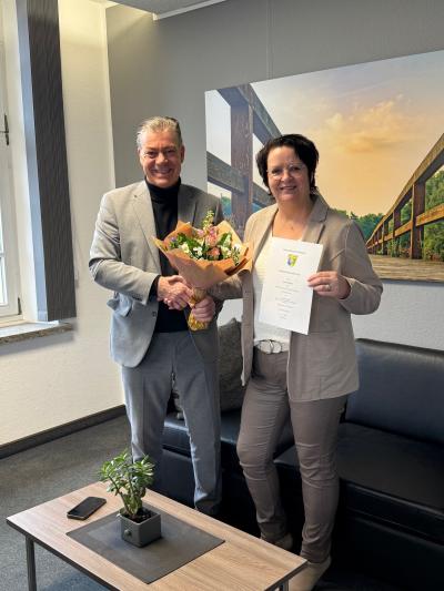 Der Bürgermeister Frank Gellen hat einen grauen Anzug an und steht links neben Frau Beckers, die ebenfalls in grau gekleidet ist. Beide geben sich die Hand und Frau Beckers hält einen Blumenstrauß in der Hand mit weißen und rosa Blüten. Frau Beckers hält zudem eine Urkunde in ihrer linken Hand. Im Vordergrund steht ein holzfarbiger Holztisch und im Hintergrund eine schwarze Couch. An der Wand hängt ein Bild mit einer grünen Landschaft.
