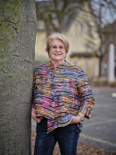 Karin Claeßen steht auf dem Kreuzherrenplatz und lehnt sich links an einem Baum an und lächelt in die Kamera. Sie hat einen blonden Kurzhaarschnitt und eine Brille. Sie trägt eine gestreifte Bluse mit vielen bunten Streifen. Im Hintergrund sieht man einen gepflasterten Platz und ein gelbes Haus.