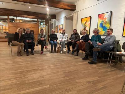 Eine Gruppe von Menschen sitzt in einem Halbkreis zusammen und schaut lächelt in die Kamera. Im Vordergrund ist ein brauner Parkettboden zu sehen und im Hintergrund Holzbalken und bunte Gemälde an der Wand. Vier Männer und fünf Frauen sitzen mittig im Bild.