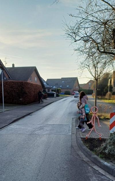 Im Vordergrund führt eine Straße in Richtung Norden. Im Hintergrund ist ein leicht bewölkter und blauer Himmel zu sehen. Mittig im Bild steht eine kleine Figur an einem Straßenübergang und reflektiert rot oranges Licht. Dahinter läuft eine Mutter mit ihrer Tochter in der Hand über die Straße.