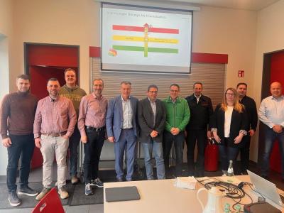 Elf Personen stehen vor einer Leinwand und gucken lächelnd in die Kamera. Auf der Leinwand sieht man oben im Bild die Abgrenzungen der Störungs- und Krisensituation in den Farben gelb, grün oder rot. Im Vordergrund ist ein Tisch mit Kabeln, Laptops und Flaschen zu sehen. Der Raum ist ausgestattet mit roten Farbakzenten. In der Mitte der Menschengruppe steht der Bürgermeister Frank Gellen und der stellvertretende Bürgermeister Dieter Dresen.