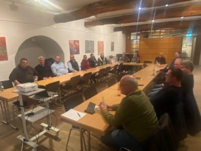 Ein Reihe von Tischen steht in U-Form im Kultursaal der Burg Brüggen. Im Hintergrund sieht man eine Holzfront und an der Wand hängen zahlreiche bunte Gemälde. Auf den tischen stehen Flaschen und es liegen Dokumente verstreut. An der Decke sind Holzbalken angebracht. Die Gruppe schaut in die Kamera.