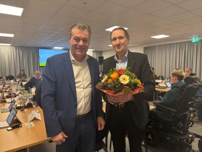 Der Bürgermeister Frank Gellen hat eine dunkel blauen Anzug an und steht neben links Berthold Bauer, der einen schwarzen Anzug trägt. Herr Bauer hält einen Blumenstrauß mit roten und gelben Blumen in den Händen. Im Hintergrund findet die Ratssitzung statt. Dafür stehen viele Tische aneinander gereiht und viele Leute sitzen dort. Auf den Tischen liegen Laptops, Tablets, Flaschen und Dokumente.