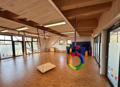 Eine große lichtdurchflutete Sporthalle mit hölzernen Boden und einer Holzdecke bestehend aus hellem und dunklen Holz ist zu sehen. Rechts und Links im Bild sieht man undurchsichtige und große Fenster mit bunten Figuren, der Sonne und Wolken darauf verziert. Im Vordergrund hängt ein regenbogenfarbener Reifen von der Decke. Daneben hängt mittig eine Plattform von der Decke. Links im Bild hängt ein großes Kletternetz von der Decke.
