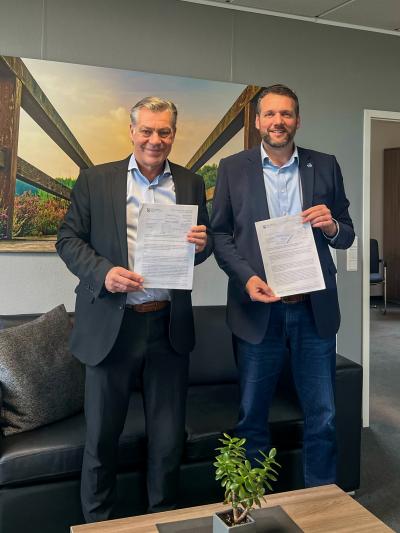 Frank Gellen und Andreas Gisbertz halten beide einen unterschriebenen Vertrag in den Händen und lächeln in die Kamera. Herr Gellen steht links im Bild und trägt einen schwarzen Anzug. Herr Gisbertz steht rechts im Bild und trägt einen dunkel blauen Anzug. Im Vordergrund steht ein holzfarbiger Tisch mit einer grünen Pflanze drauf. Im Hintergrund sieht man ein großes Bild, dass einen hölzernen Weg mit Geländer und eine grüne Naturlandschaft zeigt. Außerdem steht eine schwarze Ledercouch im Hintergrund.