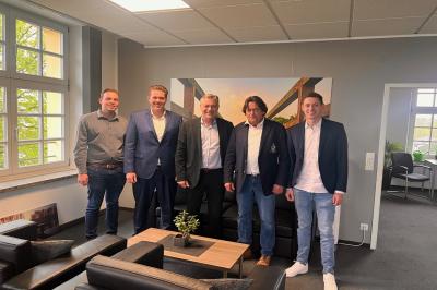 Fünf Herren stehen nebeneinander und lächeln in die Kamera. Der Herr links im Bild trägt ein dunkelgraues Hemd. Rechts neben ihm steht ein Herr in einem dunkelblauen Anzug. Mittig steht der Bürgermeister Frank Gellen in einem schwarzen Anzug. Die zwei Herren rechts im Bild tragen dunkelblaue Anzüge und blaue Jeans. Im Hintergrund sieht man eine schwarze Couch und ein Wandbild. Im Vordergrund sind zwei schwarze Sessel und ein holzfarbiger Couchtisch.