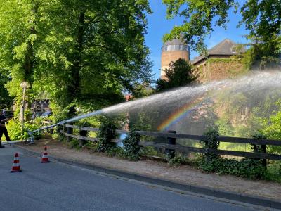 Ein Feuerwehrmann links im Bild hält einen Schlauch in den Burgweiher. Im Vordergrund ist eine asphaltierte Straße zu sehen. Im Hintergrund steht die Burg Brüggen, die links und rechts umgeben ist von grünen Bäumen. Pilonen signalsieren den Einsatz der Feuerwehr in dem Bereich. Der Himmel strahlt blau ohne Wolken.